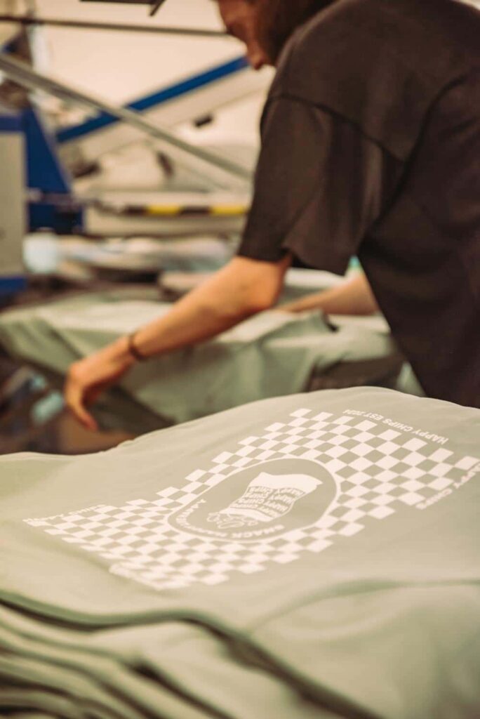 Bedruckte grüne T-Shirts liegen gestapelt vor einer Person an einer Textildruckmaschine.